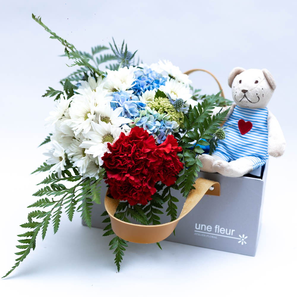 Ursinho de Pelúcia Fabler Björn Arranjo de flores com margaridas brancas, hortênsias azuis, cravos vermelhos e folhagens, acompanhado de ursinho de pelúcia com roupa listrada e coração vermelho, em caixa cinza Une Fleur dans la Ville.