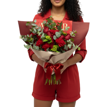 Mulher segurando um bouquet de flores vermelhas premium com roses, cravos e folhagens sofisticadas, embalado em papel bege com laço vermelho, da Une Fleur dans la Ville.