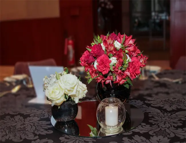 fotografia de flores vermelhas (Antúrios, Gérberas e Helicônias). O arranjo, que também tem folhas verdes, está sobre uma mesa de vidro iluminada por um lustre de cristais, em uma sala com portas de madeira e vitrais, o que exemplifica que a presença de flores e plantas pode transformar ainda mais os ambientes de trabalho.