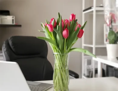 Arranjo de flores rosas com folhas verdes em vaso transparente sobre uma mesa de um escritório. Ao lado do arranjo há um laptop.