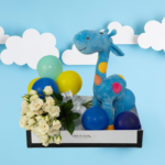 "Arranjo infantil composto por um pelúcia de girafa azul com bolinhas coloridas , mini buquê de flores brancas e pequenos balões coloridos, apresentados em uma caixa elegante."