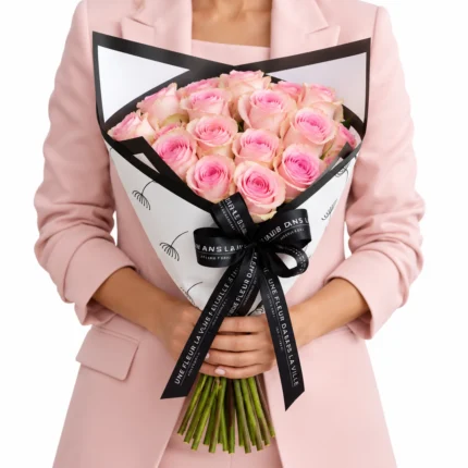 Buquê Lily Rose com rosas cor-de-rosa em tons suaves, embalagem branca com bordas pretas e laço personalizado Une Fleur Dans La Ville, segurado por mulher com blazer rosa-claro.