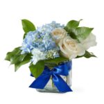 Arranjo com rosas brancas e hortênsias azuis, envolvido por um laço azul vibrante, a proposta transmite tranquilidade, elegância clássica e serenidade emocional. Ele é ideal para ocasiões mais delicadas, de agradecimento, nascimento, harmonia ou celebrações refinadas.