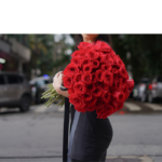 Mulher vestindo blusa vermelha segurando um bouquet de 50 rosas vermelhas com laço preto personalizado.