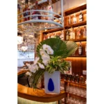 Arranjo contemporâneo com orquídeas brancas, folhagem exótica e vaso artístico de porcelana branca com detalhe azul cobalto.