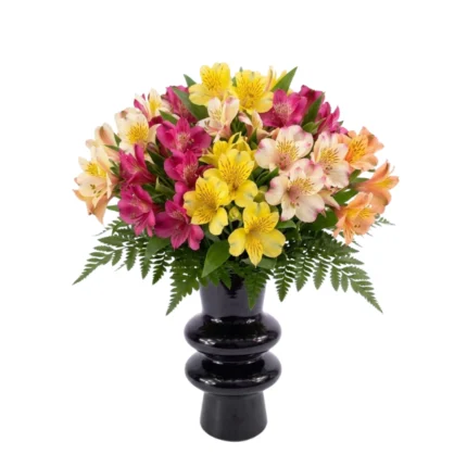 Texto Alternativo (Acessibilidade): > Arranjo de flores alstroemérias coloridas em tons de amarelo, rosa e laranja, com folhagens verdes na base, apresentado em um vaso de cerâmica preto brilhante com design de esferas empilhadas sobre fundo branco.