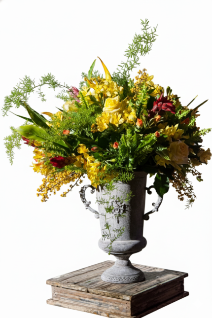 Arranjo floral tropical exuberante com flores amarelas, folhagens volumosas e detalhes em vermelho, em vaso clássico de latão tipo Ânfora com alças ornamentais, composição sofisticada e imponente.