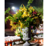 Arranjo floral tropical exuberante com flores amarelas, folhagens volumosas e detalhes em vermelho, em vaso clássico de pedra com alças ornamentais, composição sofisticada e imponente.