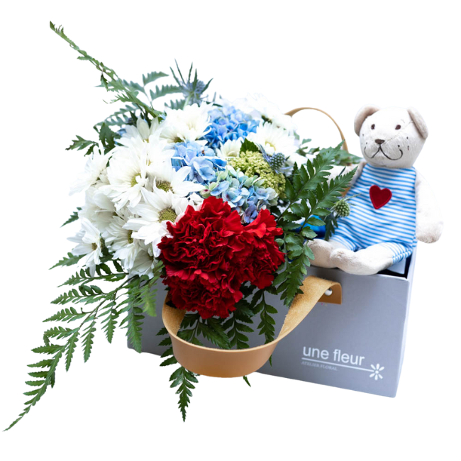 Caixa cinza Une Fleur com alça de couro com margaridas brancas, hortênsias azuis e vravos vermelhos acompanhada de ursinho de pelúcia sueco Fabler Björn.