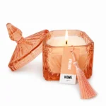 Vela aromática acesa em pote de vidro facetado âmbar/rose gold com tampa pontiaguda trabalhada e pingente de tassel em tom pêssego, fragrância Uolong Home Fragrance.